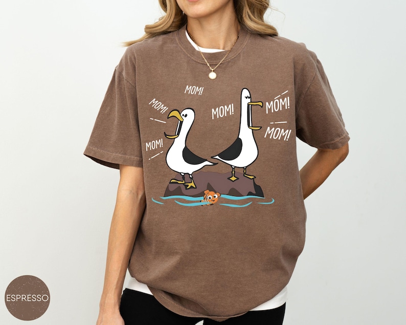 May include: Espresso-coloured t-shirt with a cartoon illustration of two seagulls on a rock, with the word "MOM!" coming from their beaks. A small orange fish is in the water below. The shirt has a relaxed fit.