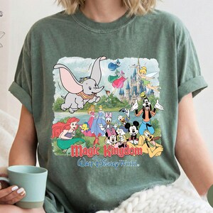 May include: A moss green short-sleeve t-shirt featuring a graphic print of various Disney characters including Dumbo, Mickey Mouse, and Ariel. The text Magic Kingdom and Walt Disney World is printed in red and blue below the illustrated characters.
