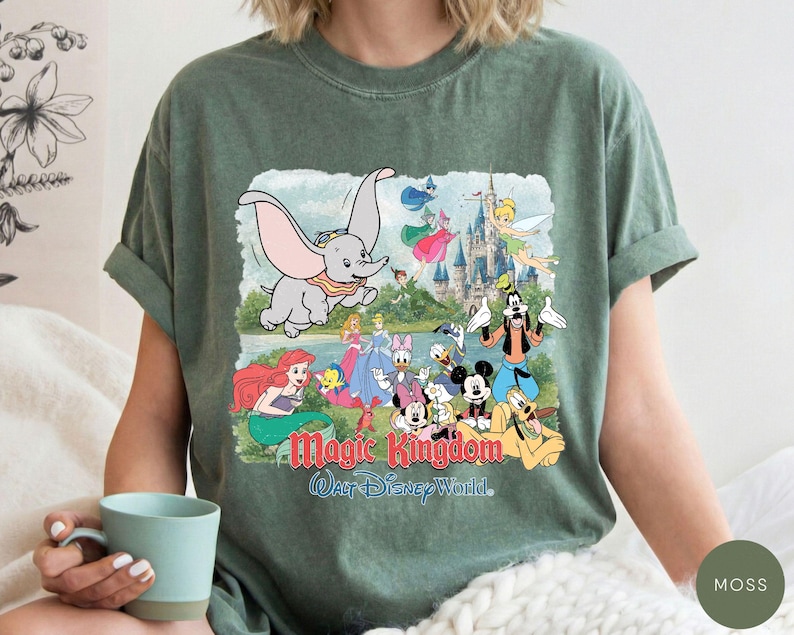 May include: A moss green short-sleeve t-shirt featuring a graphic print of various Disney characters including Dumbo, Mickey Mouse, and Ariel. The text Magic Kingdom and Walt Disney World is printed in red and blue below the illustrated characters.