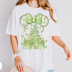 K&ouml;nnte beinhalten: Wei&szlig;es T-Shirt mit einem gr&uuml;nen Schlossdesign, einer Schleife und Mickey Mouse Ohren. Das Design beinhaltet Kleebl&auml;tter und Feuerwerk, was einen festlichen Look erzeugt. Das Shirt ist ein bequemer, l&auml;ssiger Stil.