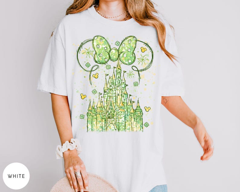 K&ouml;nnte beinhalten: Wei&szlig;es T-Shirt mit einem gr&uuml;nen Schlossdesign, einer Schleife und Mickey Mouse Ohren. Das Design beinhaltet Kleebl&auml;tter und Feuerwerk, was einen festlichen Look erzeugt. Das Shirt ist ein bequemer, l&auml;ssiger Stil.
