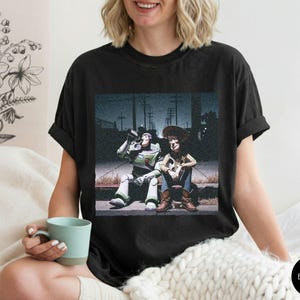 May include: A black short-sleeved t-shirt featuring a graphic print of the characters Buzz Lightyear and Woody sitting together on a curb at night. The design shows the two figures in a cinematic style against a dark, urban street background.