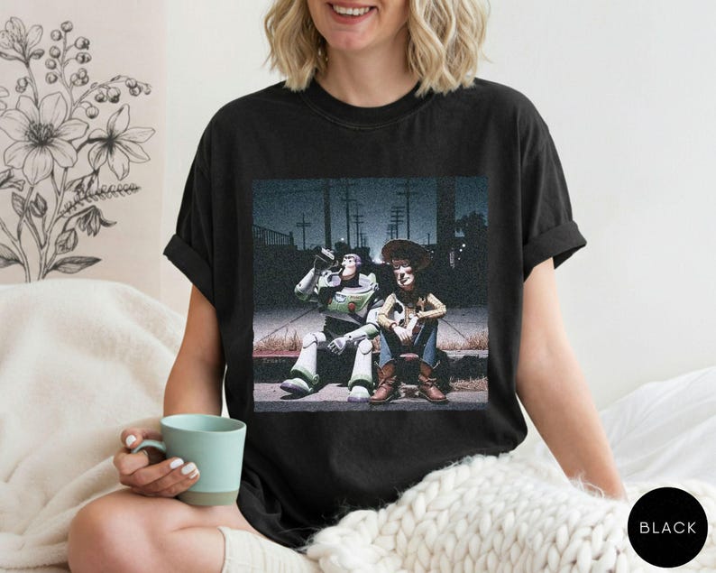 May include: A black short-sleeved t-shirt featuring a graphic print of the characters Buzz Lightyear and Woody sitting together on a curb at night. The design shows the two figures in a cinematic style against a dark, urban street background.