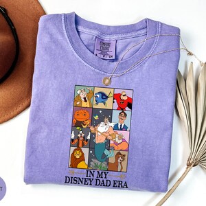 May include: A violet Comfort Colors t-shirt featuring a graphic of Disney characters in a grid. The text "IN MY DISNEY DAD ERA" is below the grid. A gold necklace and a brown hat are also in the image.