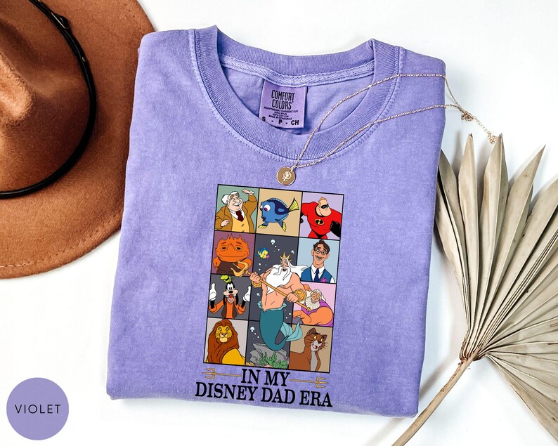 May include: A violet Comfort Colors t-shirt featuring a graphic of Disney characters in a grid. The text "IN MY DISNEY DAD ERA" is below the grid. A gold necklace and a brown hat are also in the image.