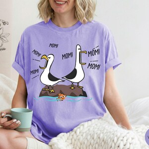 May include: Violet t-shirt with a cartoon design of two seagulls shouting "MOM!" above a small fish. The seagulls are on a rock in the sea. The shirt is made of a soft, comfortable material.