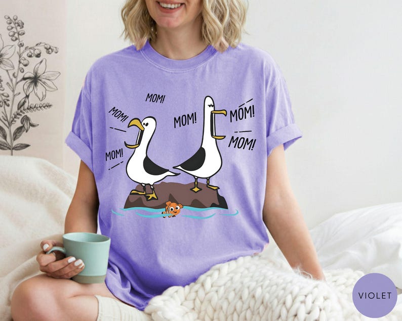 May include: Violet t-shirt with a cartoon design of two seagulls shouting "MOM!" above a small fish. The seagulls are on a rock in the sea. The shirt is made of a soft, comfortable material.