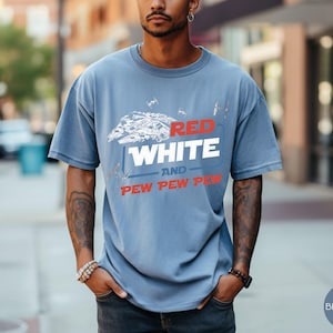 May include: A dusty blue short-sleeve t-shirt featuring a white graphic of a spaceship and smaller fighter crafts. The text reads RED in red, WHITE in white, AND in blue, and PEW PEW PEW in red. The shirt has a crew neckline and a relaxed fit.