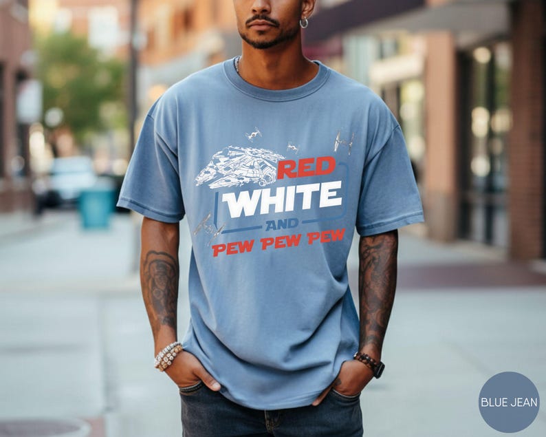 May include: A dusty blue short-sleeve t-shirt featuring a white graphic of a spaceship and smaller fighter crafts. The text reads RED in red, WHITE in white, AND in blue, and PEW PEW PEW in red. The shirt has a crew neckline and a relaxed fit.