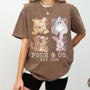 May include: Brown t-shirt featuring Winnie the Pooh, Eeyore, Tigger, and Piglet. The shirt has the text "POOH & CO. EST. 1926" in a vintage style. The shirt is a relaxed fit and has a crew neck.