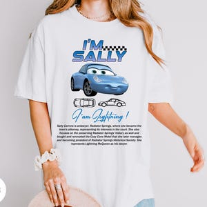 Puede incluir: Camiseta blanca con un coche de dibujos animados azul y el texto "I'M SALLY". Debajo del coche est&aacute; el texto "I'am Lightning!". La camiseta tambi&eacute;n tiene texto que describe al personaje Sally Carrera.