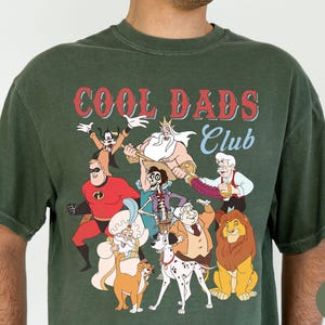 May include: Moss green t-shirt featuring a graphic of various cartoon characters with the text "COOL DADS Club" in red and white. The design includes characters like Mr. Incredible, Simba, and Goofy.