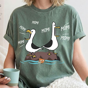 May include: Green t-shirt with a cartoon design of two seagulls, each with the word "MOM!" above them. A small orange fish swims in the water below. The shirt is a relaxed fit, suitable for everyday wear.