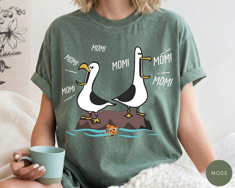 May include: Green t-shirt with a cartoon design of two seagulls, each with the word "MOM!" above them. A small orange fish swims in the water below. The shirt is a relaxed fit, suitable for everyday wear.