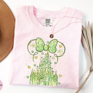 K&ouml;nnte beinhalten: Ein hellrosa T-Shirt mit einer gr&uuml;n-gelben Grafik eines Schlosses und einer Schleife. Das Shirt hat einen Rundhalsausschnitt und kurze &Auml;rmel. Eine goldene Halskette ist &uuml;ber das Shirt drapiert. Das Shirt ist auf einer wei&szlig;en Oberfl&auml;che gefaltet.