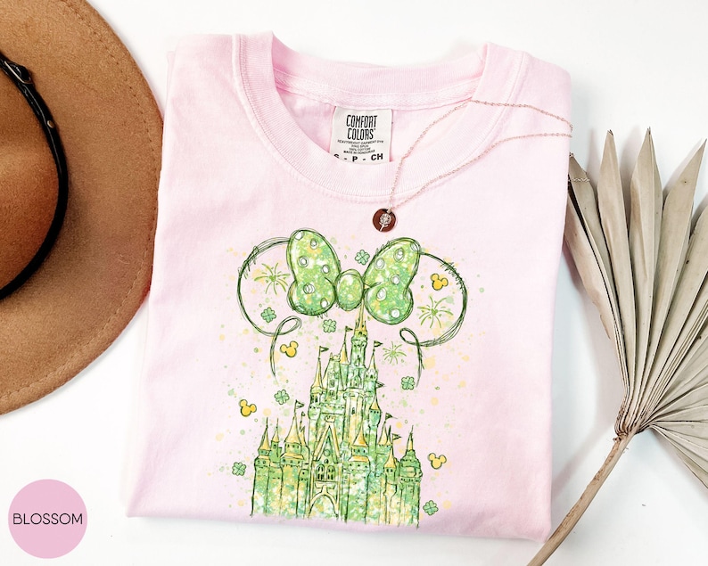 K&ouml;nnte beinhalten: Ein hellrosa T-Shirt mit einer gr&uuml;n-gelben Grafik eines Schlosses und einer Schleife. Das Shirt hat einen Rundhalsausschnitt und kurze &Auml;rmel. Eine goldene Halskette ist &uuml;ber das Shirt drapiert. Das Shirt ist auf einer wei&szlig;en Oberfl&auml;che gefaltet.