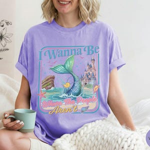 May include: Violet t-shirt with a mermaid graphic. The design features a mermaid tail, a castle, and the text "I Wanna Be Where The People Aren't". The shirt is a comfortable, casual style.
