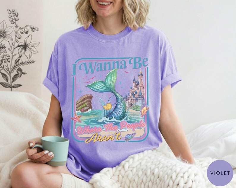 May include: Violet t-shirt with a mermaid graphic. The design features a mermaid tail, a castle, and the text "I Wanna Be Where The People Aren't". The shirt is a comfortable, casual style.