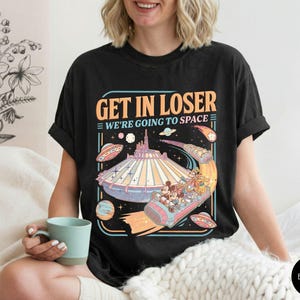 Pode incluir: Camiseta preta com um gr&aacute;fico retr&ocirc; apresentando um passeio de parque tem&aacute;tico com tema espacial e o texto "GET IN LOSER WE'RE GOING TO SPACE". O design inclui personagens de desenhos animados, planetas e discos voadores.
