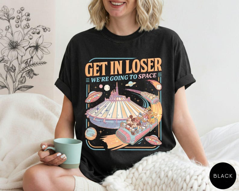 Pode incluir: Camiseta preta com um gr&aacute;fico retr&ocirc; apresentando um passeio de parque tem&aacute;tico com tema espacial e o texto "GET IN LOSER WE'RE GOING TO SPACE". O design inclui personagens de desenhos animados, planetas e discos voadores.
