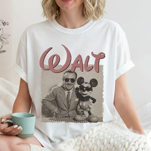 Puede incluir: Camiseta blanca con una imagen retro de Walt Disney y Mickey Mouse con gafas de sol. La palabra "Walt" est&aacute; impresa encima de la imagen en una fuente rosa vintage. La camiseta es de estilo cuello redondo cl&aacute;sico.