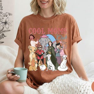 May include: A brown t-shirt with the text "COOL MOMS Club" in pink and blue. The shirt features a graphic of various cartoon characters. The person is holding a blue mug.