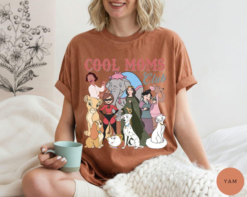 May include: A brown t-shirt with the text "COOL MOMS Club" in pink and blue. The shirt features a graphic of various cartoon characters. The person is holding a blue mug.