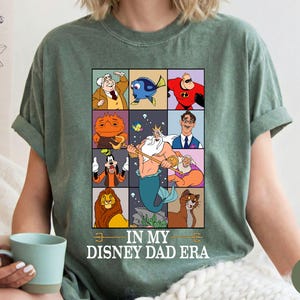 May include: Moss green t-shirt featuring a grid of Disney character illustrations. The shirt includes characters like Dory, Mr. Incredible, and King Triton. Text on the shirt reads "IN MY DISNEY DAD ERA."