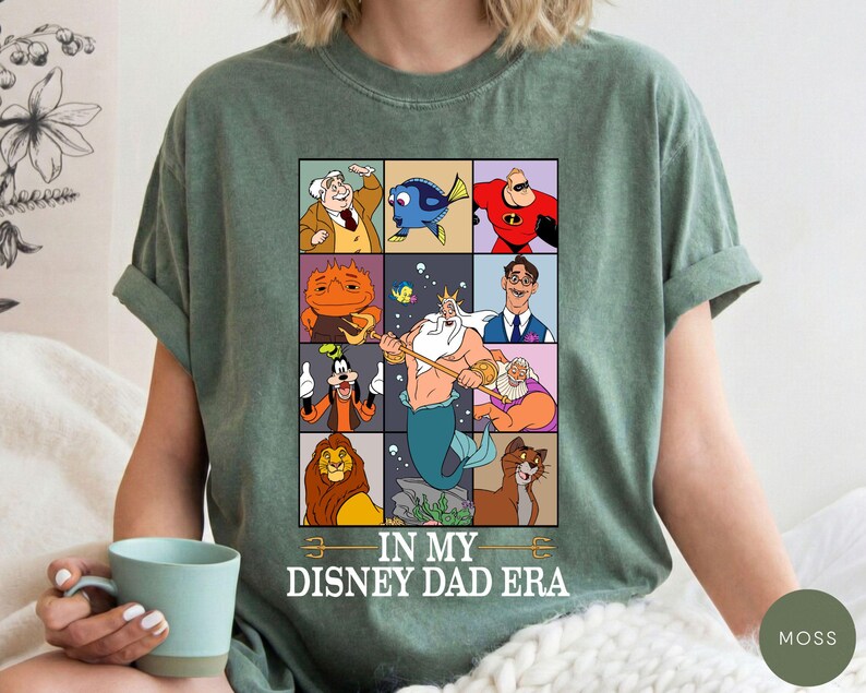 May include: Moss green t-shirt featuring a grid of Disney character illustrations. The shirt includes characters like Dory, Mr. Incredible, and King Triton. Text on the shirt reads "IN MY DISNEY DAD ERA."