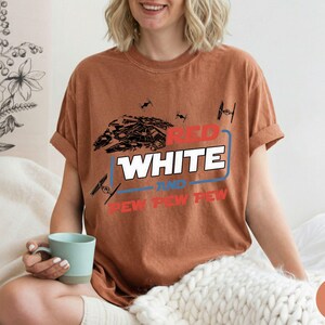 May include: A rust-colored short-sleeve t-shirt featuring a graphic print of a spaceship and TIE fighters. The text reads RED WHITE AND PEW PEW PEW in a mix of red, white, and blue colors, with a blue rectangular outline surrounding the word WHITE.