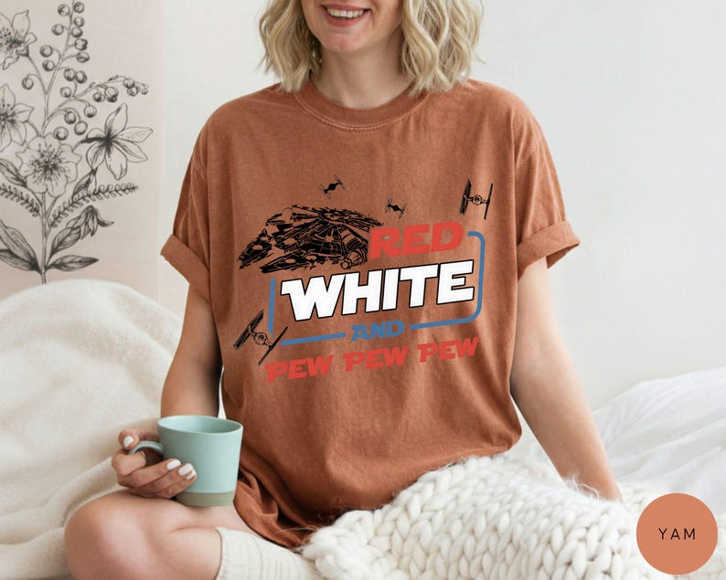 May include: A rust-colored short-sleeve t-shirt featuring a graphic print of a spaceship and TIE fighters. The text reads RED WHITE AND PEW PEW PEW in a mix of red, white, and blue colors, with a blue rectangular outline surrounding the word WHITE.
