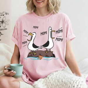 May include: Pale pink t-shirt with a cartoon design of two seagulls shouting "MOM!". The seagulls are on a rock above the water, with an orange fish below. The shirt is a light pink colour. The text "MOM!" is repeated around the seagulls.