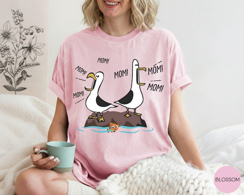 May include: Pale pink t-shirt with a cartoon design of two seagulls shouting "MOM!". The seagulls are on a rock above the water, with an orange fish below. The shirt is a light pink colour. The text "MOM!" is repeated around the seagulls.