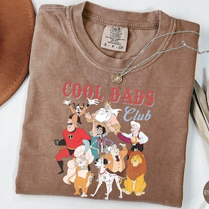 May include: Espresso-colored t-shirt with the text "COOL DADS Club" and cartoon characters. The shirt has a crew neck and short sleeves. A silver necklace with a pendant is draped across the shirt. The shirt is size S-P-CH.