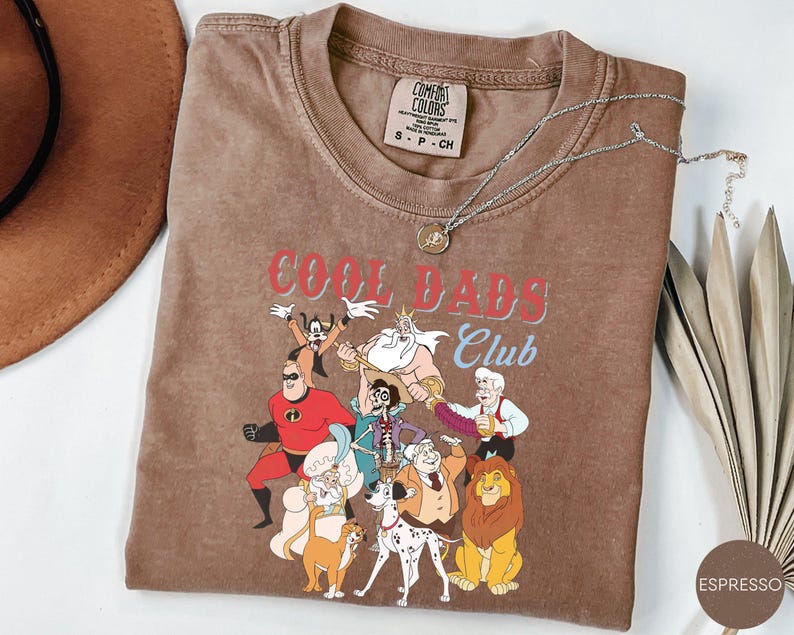 May include: Espresso-colored t-shirt with the text "COOL DADS Club" and cartoon characters. The shirt has a crew neck and short sleeves. A silver necklace with a pendant is draped across the shirt. The shirt is size S-P-CH.