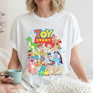 May include: A white t-shirt featuring a colourful graphic print of Toy Story characters including Woody, Buzz Lightyear, Jessie, Rex, and others. The design includes the Toy Story logo in red and yellow, surrounded by scribbled crayon-style drawings.