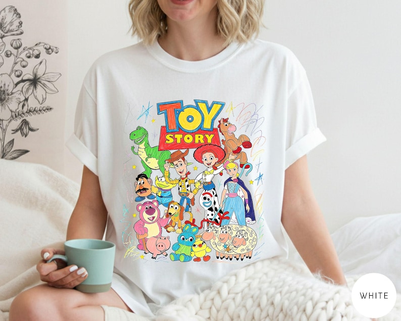 May include: A white t-shirt featuring a colourful graphic print of Toy Story characters including Woody, Buzz Lightyear, Jessie, Rex, and others. The design includes the Toy Story logo in red and yellow, surrounded by scribbled crayon-style drawings.