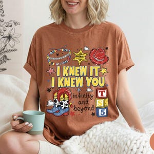 May include: A rust-colored short-sleeve t-shirt featuring a graphic print with the text I KNEW IT I KNEW YOU infinity and beyond. The design includes a sheriff badge, a red cowboy hat, colorful beaded bracelets, cowboy boots, and toy blocks.