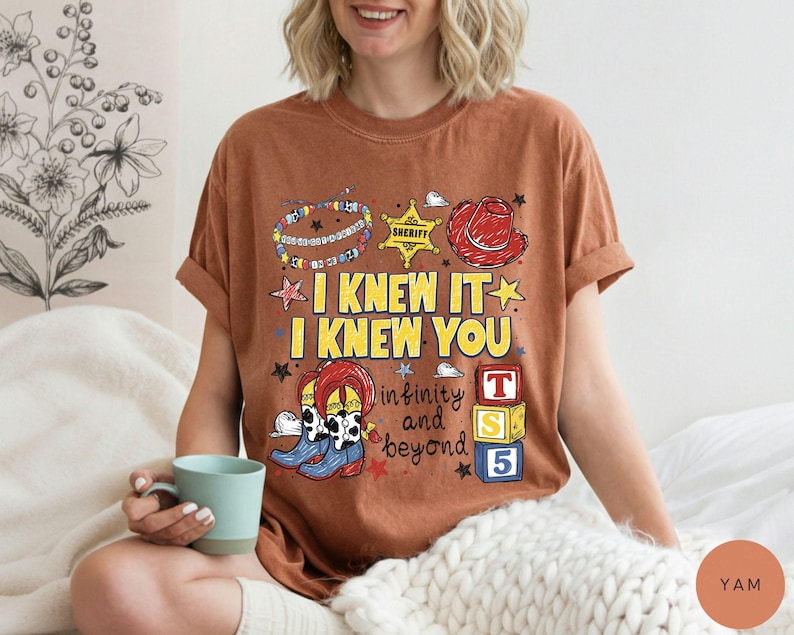 May include: A rust-colored short-sleeve t-shirt featuring a graphic print with the text I KNEW IT I KNEW YOU infinity and beyond. The design includes a sheriff badge, a red cowboy hat, colorful beaded bracelets, cowboy boots, and toy blocks.