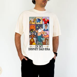 May include: Ivory-colored t-shirt featuring a grid of Disney character illustrations. The shirt includes characters like Dory, Mr. Incredible, and Simba. Text on the shirt reads "IN MY DISNEY DAD ERA."