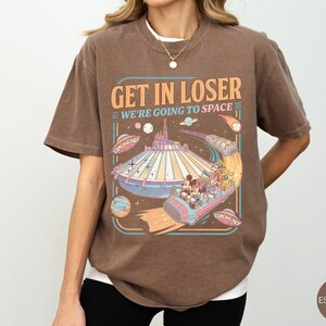 Pode incluir: Camiseta marrom com um gr&aacute;fico de um passeio de parque tem&aacute;tico com tema espacial. O texto "GET IN LOSER WE'RE GOING TO SPACE" &eacute; exibido acima do passeio. O design inclui personagens de desenhos animados e discos voadores.