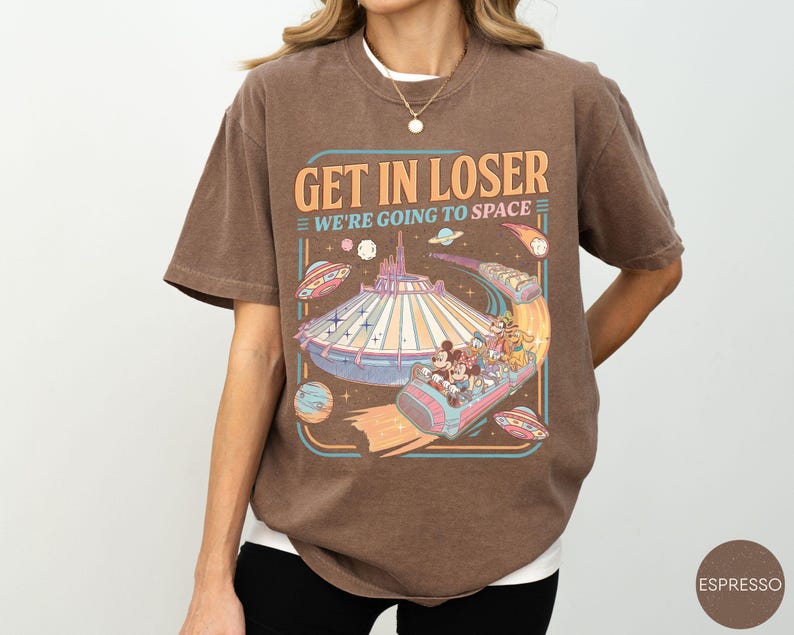 Pode incluir: Camiseta marrom com um gr&aacute;fico de um passeio de parque tem&aacute;tico com tema espacial. O texto "GET IN LOSER WE'RE GOING TO SPACE" &eacute; exibido acima do passeio. O design inclui personagens de desenhos animados e discos voadores.