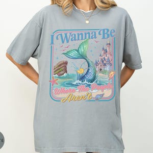 May include: A light gray t-shirt with a graphic of a mermaid tail emerging from the ocean, with a castle in the background. The text "I Wanna Be Where The People Aren't" is displayed in a retro font.