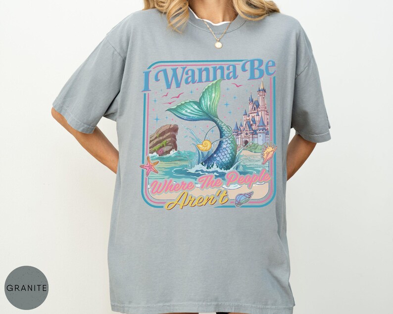 May include: A light gray t-shirt with a graphic of a mermaid tail emerging from the ocean, with a castle in the background. The text "I Wanna Be Where The People Aren't" is displayed in a retro font.