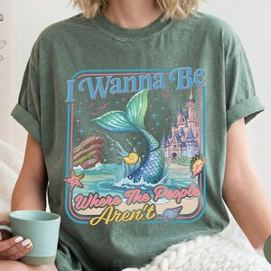 May include: Moss green t-shirt featuring a mermaid tail design with the text "I Wanna Be Where The People Aren't". The graphic includes a mermaid tail, castle, and ocean elements in a retro style.