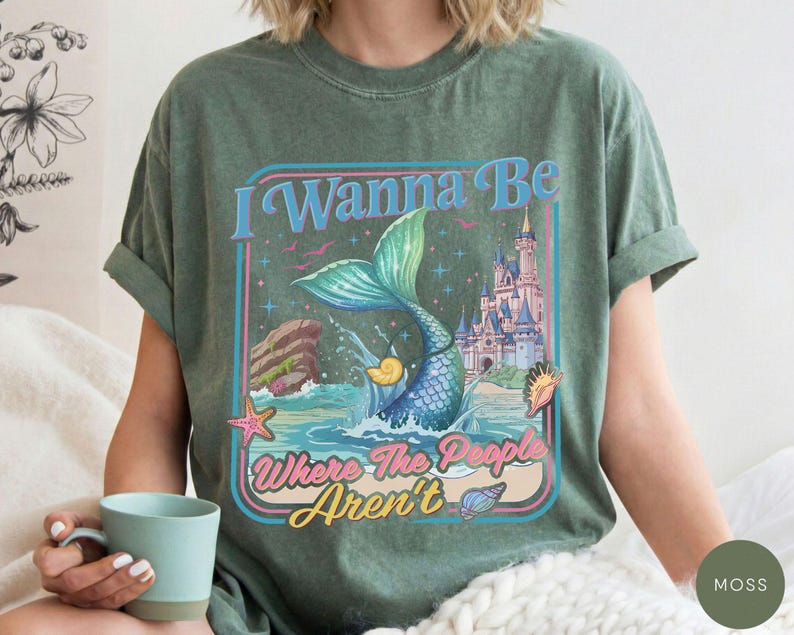 May include: Moss green t-shirt featuring a mermaid tail design with the text "I Wanna Be Where The People Aren't". The graphic includes a mermaid tail, castle, and ocean elements in a retro style.