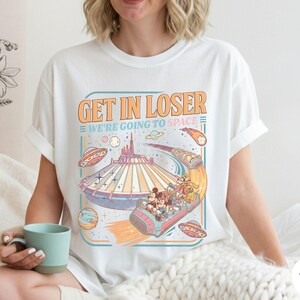 Pode incluir: Camiseta branca com um gr&aacute;fico de estilo retr&ocirc; com o texto "GET IN LOSER WE'RE GOING TO SPACE". O design inclui uma ilustra&ccedil;&atilde;o com tema espacial com uma montanha-russa, planetas e personagens de desenhos animados. A camisa &eacute; um estilo casual e unissex.