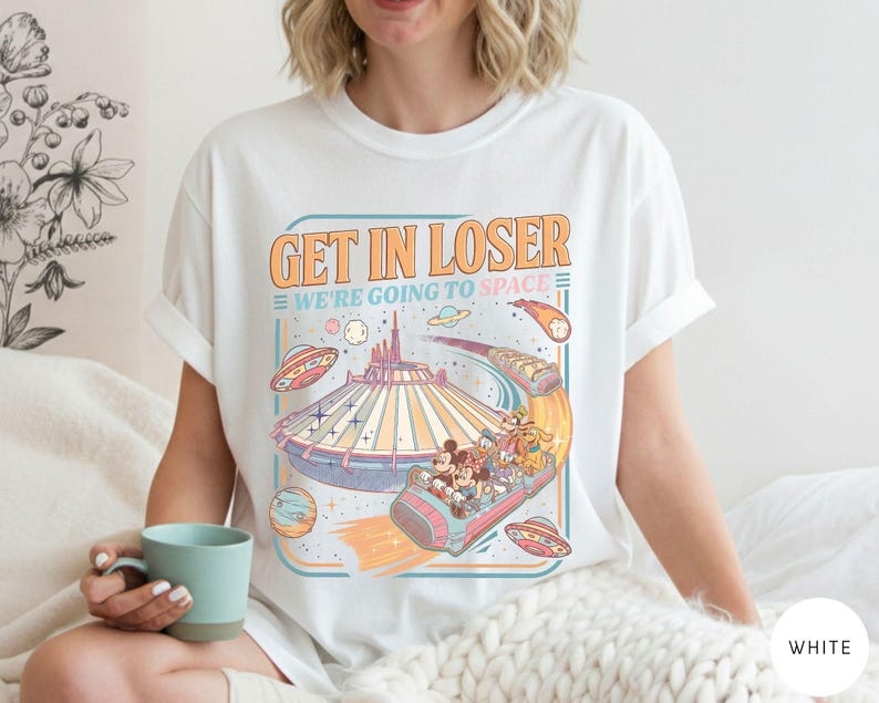Pode incluir: Camiseta branca com um gr&aacute;fico de estilo retr&ocirc; com o texto "GET IN LOSER WE'RE GOING TO SPACE". O design inclui uma ilustra&ccedil;&atilde;o com tema espacial com uma montanha-russa, planetas e personagens de desenhos animados. A camisa &eacute; um estilo casual e unissex.