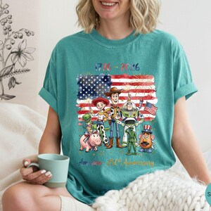 Pu&ograve; includere: T-shirt color verde acqua con grafica dei personaggi di Toy Story davanti a una bandiera americana con fuochi d'artificio. Il testo recita "1776-2016" e "America 250th Anniversary". La maglietta &egrave; in stile casual e confortevole.