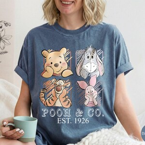 May include: Blue jean t-shirt featuring Winnie the Pooh and friends. The design includes Pooh Bear, Eeyore, Tigger, and Piglet, with the text "POOH & CO. EST. 1926". The shirt has rolled-up sleeves.